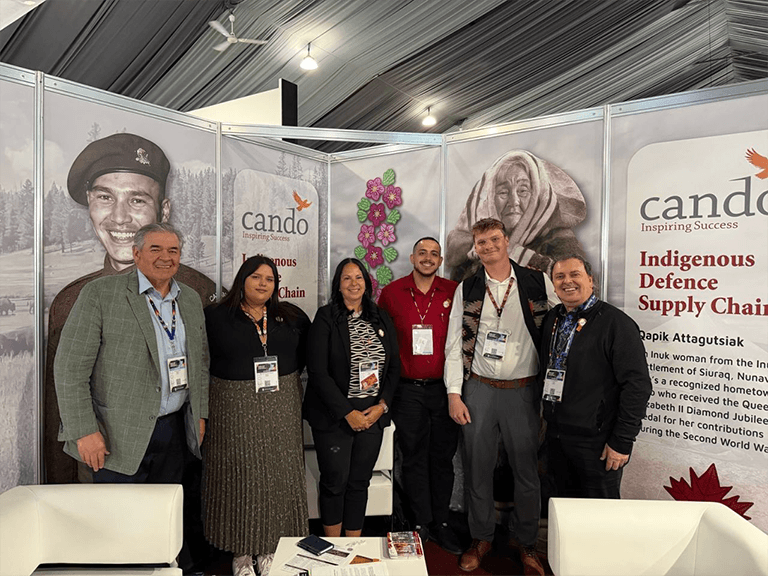 Photo of six adults standing in front of the Cando CANSEC pavilion featuring Indigenous war heroes.