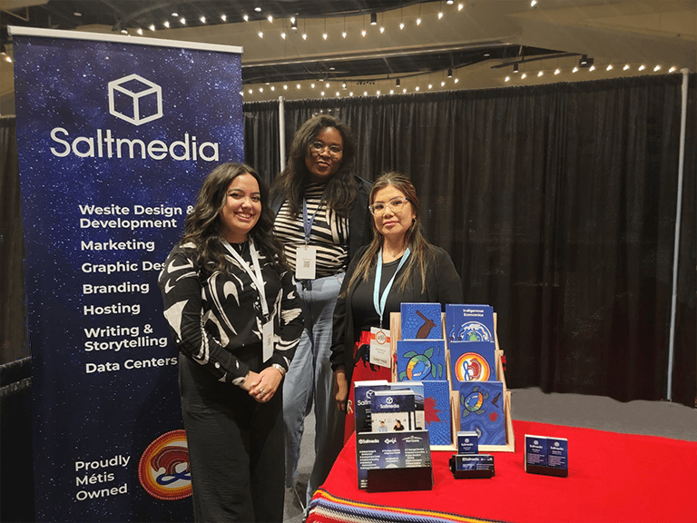 Natalya, Brandi, and Ellie stand at the Saltmedia booth at Trailblazers featuring a large Saltmedia pullup banner and display. Everyone is wearing black t-shirts with the Saltmedia 15-year anniversary logo.